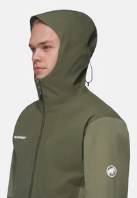 Grøn vandtæt jakke med høj krave, justerbar hætte, sidelomme med zip og hvidt logo på brystet og ærmet. Glat tekstur.