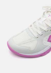 Chaussure de sport blanche avec tige en mesh, accents de semelle rose et design texturé. Présente un système de laçage allongé et une languette rembourrée.