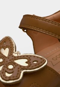 Καφέ συνθετικό σανδάλι με διακοσμητικό στρώσεων σε σχήμα χρυσής πεταλούδας, με λεία υφή και ρυθμιζόμενο λουράκι για άνετη εφαρμογή.