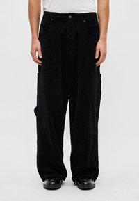 Fekete kordbársony nadrág laza fazonban, két első zsebbel és egy oldalzsebbel, díszített texturált szövettel és gombos záródással.