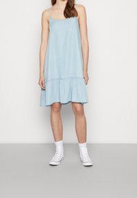 Svetle modré bezrukávové šaty s vrstveným volánkovým lemem. Vyrobené z měkké tkaniny; v kombinaci s bílými vysokými teniskami a kotníkovými ponožkami.