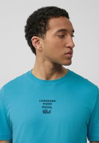 Turquoise katoenen t-shirt met een ronde halslijn en korte mouwen. Bevat zwart geborduurde tekst: "LONESOME RIDER SOCIAL Club" gecentreerd op de borst.