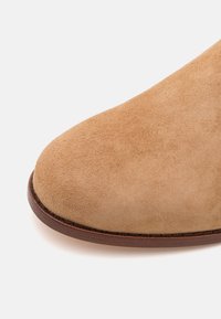 Ljusbrun mocka känga med rund tå, kontrasterad av en mörkbrun lädersula och diskret sömnad längs kanten. Slät textur.