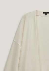 Kardigan w kolorze kremowym, zapinany z przodu, z opadającym szwem ramienia i czarną metką marki wewnątrz kołnierza.