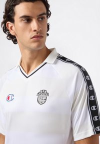 Jonge man met donker krullend haar, gekleed in een wit poloshirt met zwarte logopatches en gestreepte zwarte patronen op de mouwen.