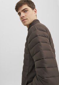 Brun puffjacka med kuddade texturer, hög krage och synlig sömnad. Lättviktsdesign med en åtsittande passform och minimalistisk stil.