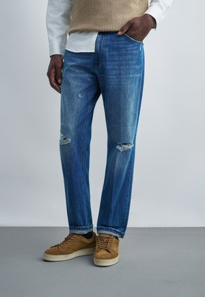 Man draagt blauwe versleten jeans, beige sneakers, wit overhemd en beige gebreid vest, staat met één hand in de zak.