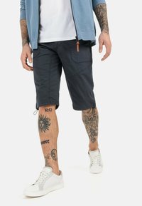 Marinefarbene Cargo-Shorts mit mehreren Taschen, aus strapazierfähigem Stoff. Tragen mit weißen Sneakers. Sichtbare Tattoos an den Beinen.