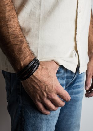 Bratara din piele neagră, cu trei benzi, având texturi netede și împletite. Puritată pe un încheietură cu păr de braț vizibil. Cămașă de culoare deschisă și blugi.
