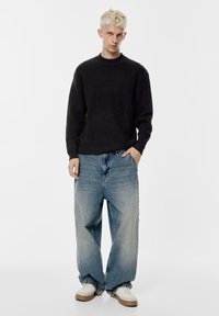 Maglione nero a coste, jeans oversize larghi blu chiaro, sneakers bianche con accenti marroni. Il modello è in piedi con una mano in tasca.