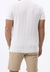 Mand iført en hvidmønstret kortærmet strikbluse og beige shorts, stående med ryggen til kameraet mod en hvid baggrund.