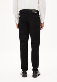 Zwarte denim jeans met een rechte pijp, vijf zakken en een logo flap op de tailleband; gedragen met zwarte schoenen en een wit shirt.