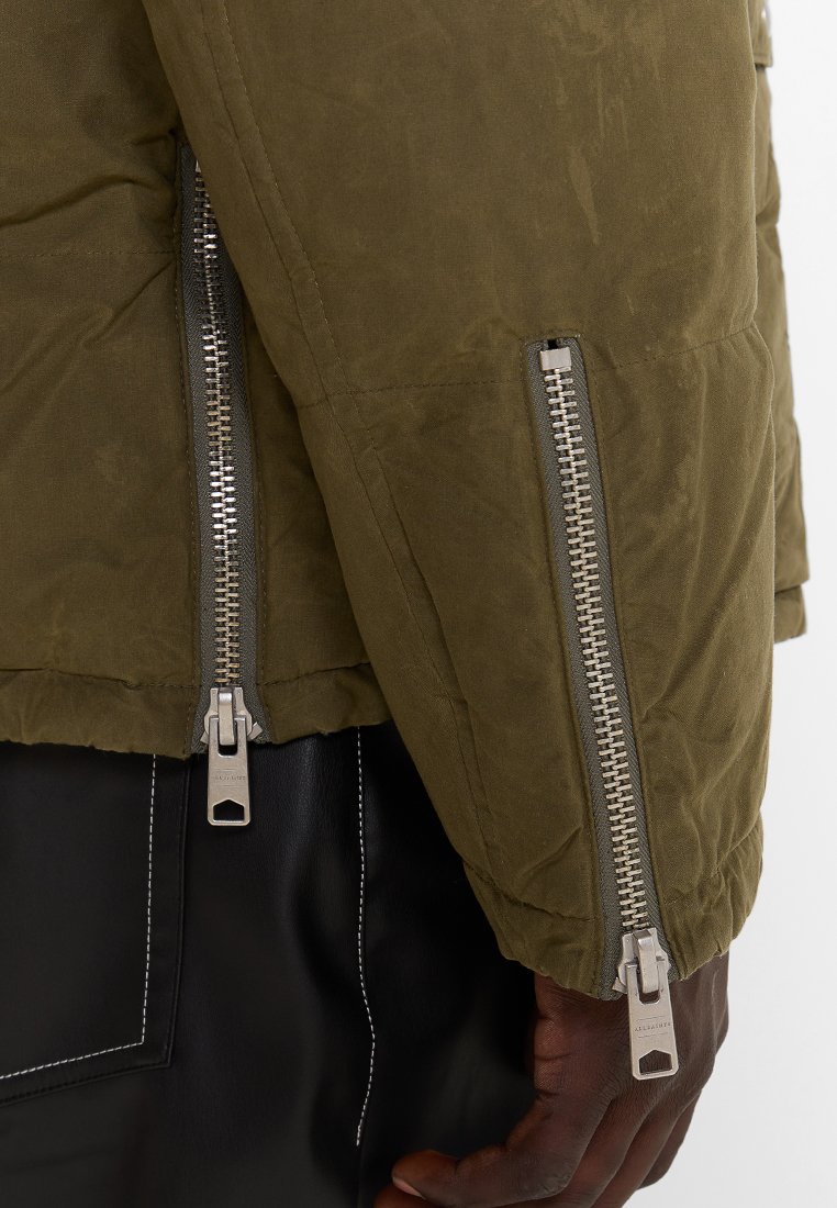 Zbliżenie na osobę w oliwkowej zielonej kurtce z dwoma srebrnymi zamkami na rękawach oraz czarnych spodniach z białymi szwami.