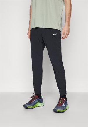 Person iført sorte Nike træningsbukser og flerfarvede sportssko står på en lysegrå gulv mod en hvid væg.