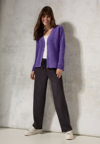 Lila texturerad kofta med ribbade ärmar, knäppning framtill, i kombination med svarta byxor med dragsko och vita sneakers.