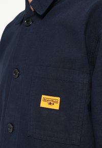 Μπλε σκούρο επαγγελματικό πουκάμισο από υφή υφάσματος, με γιακά με μύτες, δύο μαύρα κουμπιά και αριστερή τσέπη στο στήθος με κίτρινη ετικέτα.