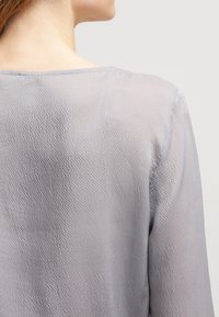 Blusa grigia chiaro, texturizzata, con una vestibilità morbida, caratterizzata da un'ampia scollatura e maniche lunghe. Il tessuto sembra morbido e leggermente trasparente.