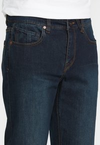 Pantalones vaqueros de mezclilla azul oscuro que cuentan con un diseño clásico de cinco bolsillos, remaches de cobre y costuras en naranja contrastante a lo largo de las costuras y la cintura.