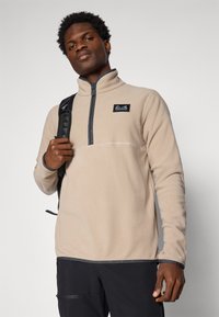 Pullover in pile beige con colletto alto, mezza zip e patch logo nero. Caratterizzato da rifiniture scure a contrasto e tessuto testurizzato. Cinghia dello zaino nera visibile.