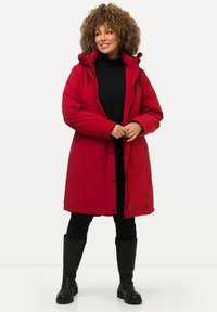 Red, knee-length parka with a hood, zippered front, and two side pockets. Black turtleneck underneath and black pants, worn with tall black boots.