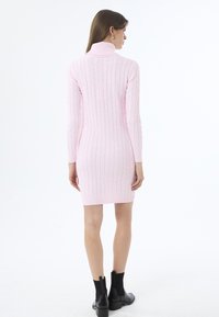 Růžové pletené šaty s copánkovým vzorem, s vysokým límcem a dlouhými rukávy. Šaty jsou přiléhavé a končí nad polovinou stehna.