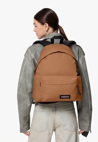 Zaino in tessuto arancione con parte superiore piatta, tasca esterna e accessori neri. Le cinghie sono regolabili e si indossa sulla schiena.
