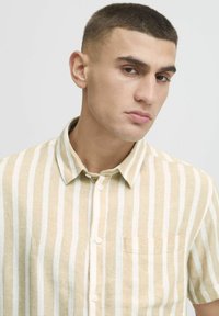 Camisa de manga corta en beige con rayas verticales blancas, hecha de una tela ligera. Presenta un cierre de botones en el frente y un bolsillo en el lado izquierdo del pecho.