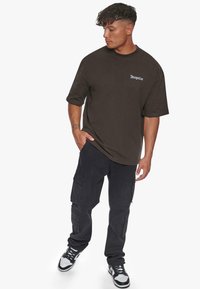 Mørkebrun oversized t-skjorte med en liten hvit logo, kombinert med svarte cargo-bukser med sidelommer og designet for uformell bruk.