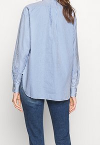 Striped blue and white button-up shirt, featuring long sleeves, a relaxed fit, side slits, and a back yoke detail. Paired with dark blue jeans.