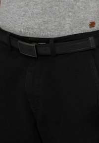 Czarne bawełniane spodnie o gładkiej fakturze, z czarnym paskiem z prostokątną klamrą. Szary sweter z dzianiny widoczny nad paskiem.