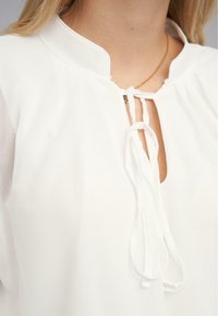 Hvid bluse med mandarin-krave, snorelukning ved halsen og gennemsigtig stof med en glat tekstur.