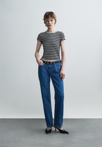 Άτομο που στέκεται με ριγέ μαύρο-λευκό κοντομάνικο τοπ, τζιν μπλε με μαύρη ζώνη και μαύρα μυτερά τακούνια σε γκρι πάτωμα.