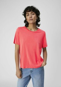 Koraal kleur t-shirt met korte mouwen, ronde halslijn, gladde stof, losse pasvorm, gecombineerd met lichtblauwe denim jeans, model met krullend haar.