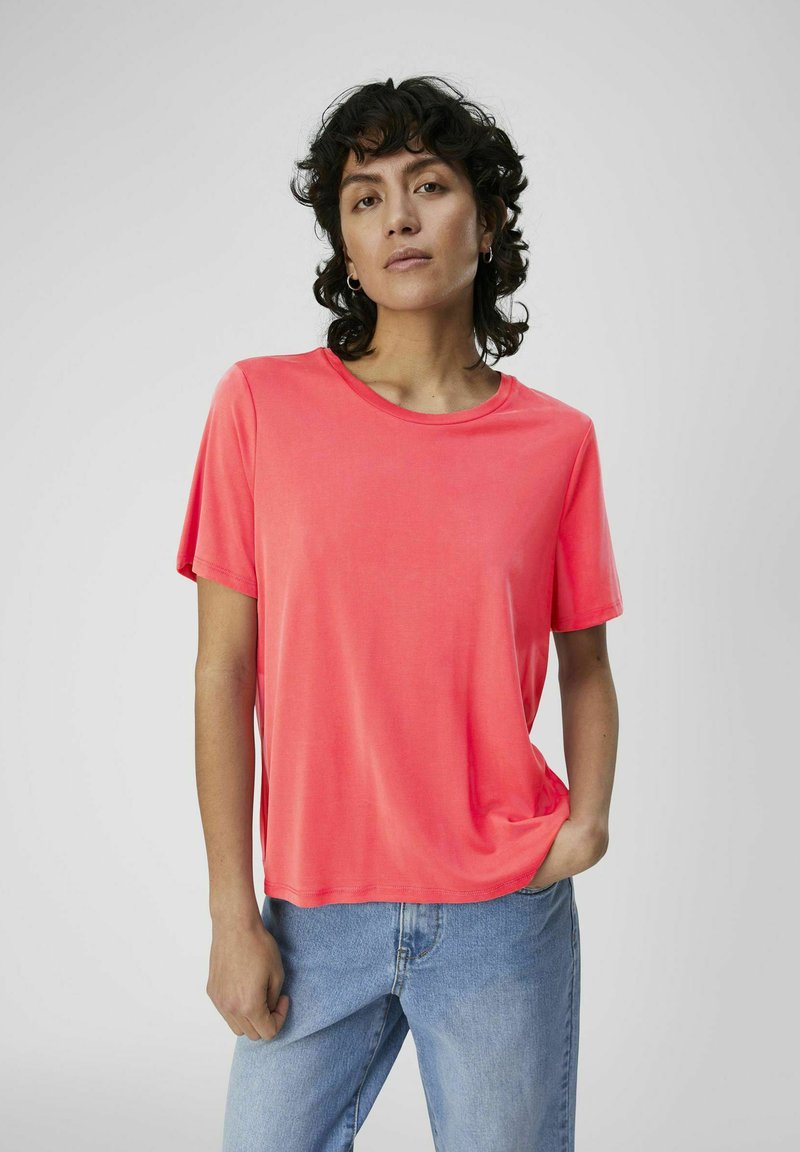 Koraal kleur t-shirt met korte mouwen, ronde halslijn, gladde stof, losse pasvorm, gecombineerd met lichtblauwe denim jeans, model met krullend haar.