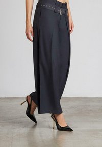 Pantalones de pierna ancha oscuros de talle alto y entallados, con un cinturón que presenta ojales de metal, combinados con tacones finos negros y brillantes.