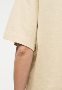 Λεπτομέρεια υφάσματος μπεζ πουκαμίσου με κοντά μανίκια που δείχνει το τελείωμα του μανικιού και τον γυμνό, λυγισμένο αγκώνα και πήχη ενός ατόμου πάνω σε απλό φόντο.