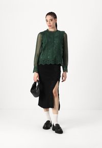 Blusa verde in pizzo con bordi arricciati e maniche lunghe, abbinata a una gonna nera con uno spacco laterale e bordo in pizzo. Borsa e scarpe nere.
