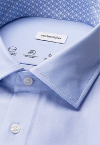 Colletto di camicia azzurro chiaro con bottoni bianchi, colletto interno a fantasia e etichetta del marchio "seidensticker" visibile all'interno.