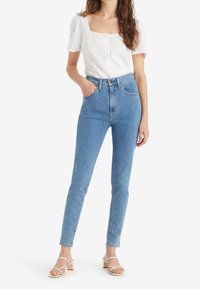 Mujer con blusa blanca de manga corta abotonada, jeans ajustados de tiro alto en azul claro y sandalias de tacón abiertas en los dedos de los pies de color blanco.