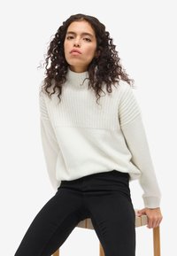 Svetra svetle barve z rebrasto pletenino, visokim ovratnikom in sproščenim krojem, v kombinaciji z črnimi ozkimi kavbojkami. Manekenka sedi na lesenem stolu.