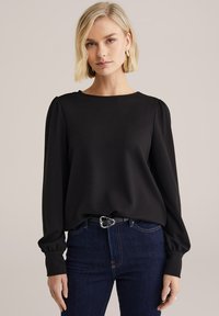 Szőke nő, fekete, texturált, hosszú ujjú blúzt visel, melynek vállai puffadtak, sötétkék farmert és fekete övet ezüst csattal.