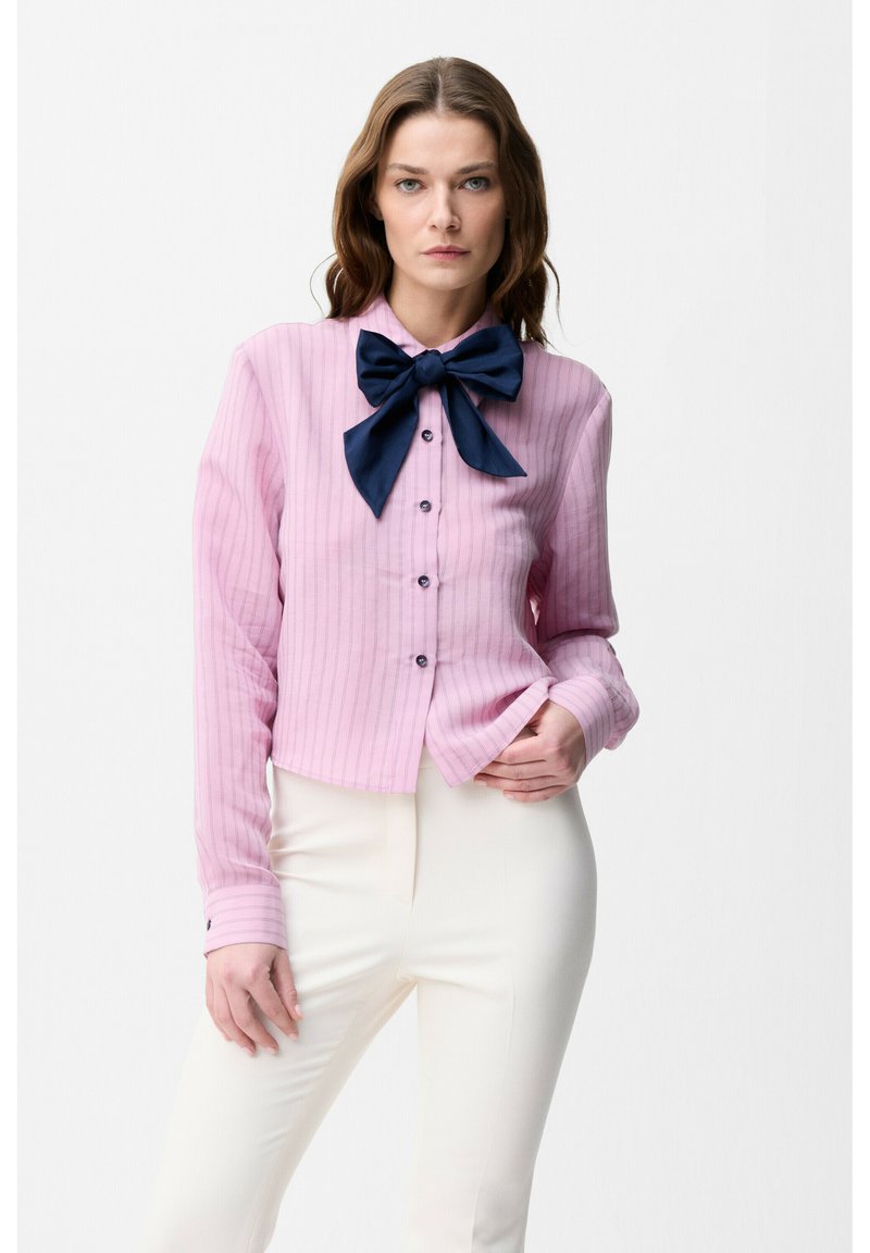 Woman with long brown hair wearing a pink striped blouse with a large navy blue bow and white pants, posing against a white background.