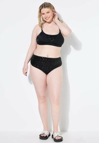 Bikini nero due pezzi con top accentato da paillettes e bottom a vita alta. La modella indossa sandali bianchi a ciabatta. Sfondo minimalista.