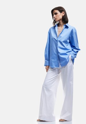 Camisa azul claro de botones con un ajuste relajado y mangas largas, combinada con pantalones anchos blancos y sandalias nude. Textura suave.
