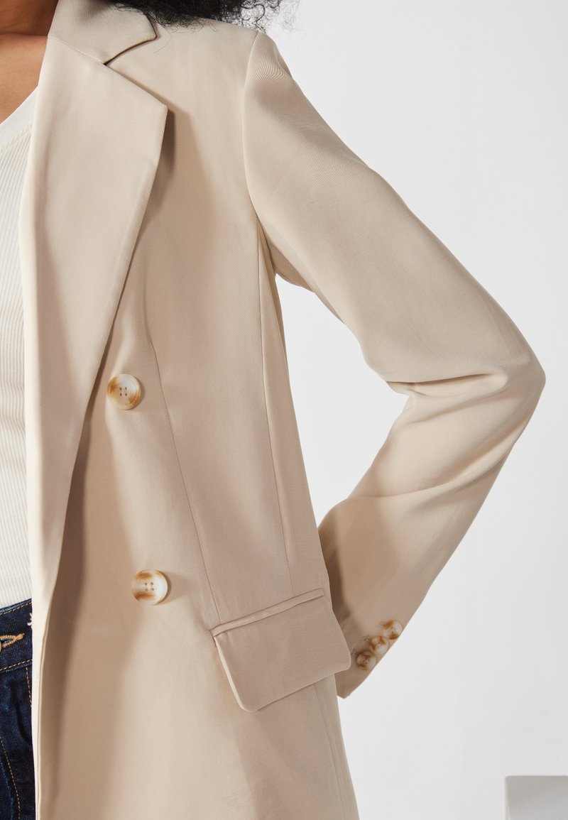 Blazer beige con una vestibilità su misura, design doppiopetto, tessuto morbido e grandi bottoni tondi. Presenta un rever a punta e spalle strutturate.