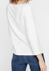 Maglietta bianca a maniche lunghe con texture e motivo a coste, scollo rotondo e vestibilità aderente, abbinata a jeans blu scuro con dettagli sulle tasche.