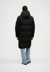 Čierna zateplená bunda s kapucňou, ktorá má dlhý strih a horizontálny prešívaný vzor, v kombinácii so svetlomodrými džínsami a čiernou obuvou.