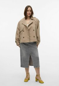 Manteau trench court beige avec des boutons à double boutonnage, associé à une jupe en denim grise avec une fente devant, et des chaussures à talons jaunes.
