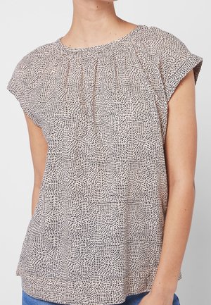 Kobieta w beżowej bluzce z krótkim rękawem, z czarnym wzorem cienkich szwów i marszczonym dekoltem, w połączeniu z niebieskimi dżinsami.