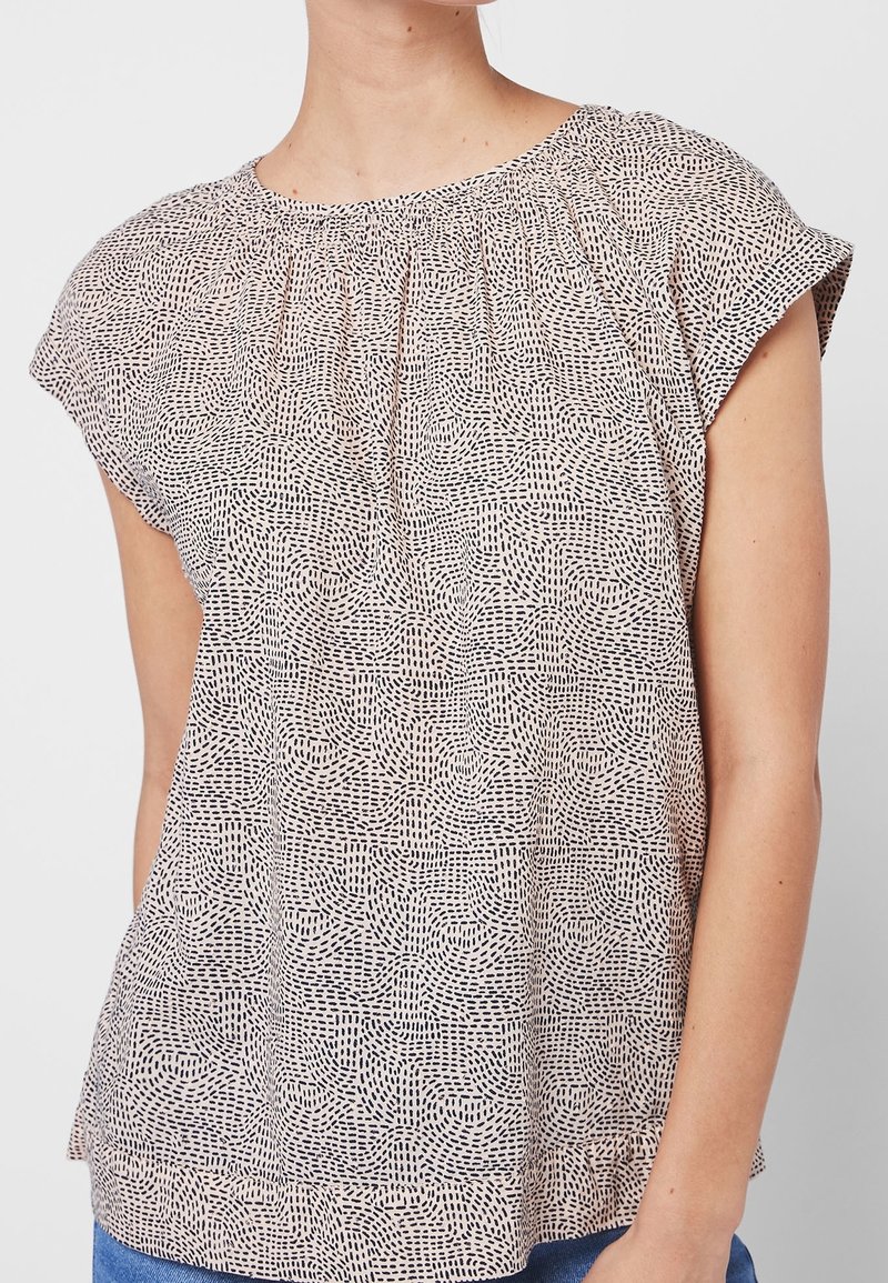 Femme portant un chemisier beige à manches courtes avec un motif de tirets noirs cousus et un col froncé, associé à un jean bleu.